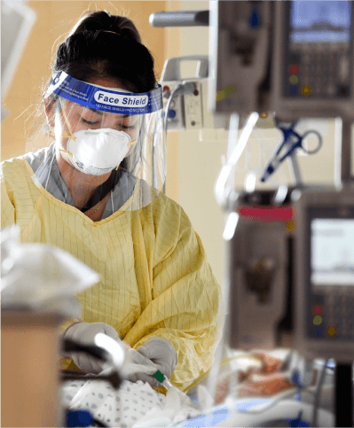 A health care worker wearing a face shield, mask, and PPE, provides care to a patient in a hospital bed. 